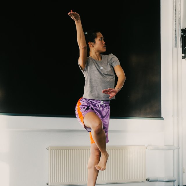 Eine performende Person balanciert auf einem Bein in einem hellen Raum. Ein Arm ist nach oben gef&uuml;hrt, der andere vor dem K&ouml;rper positioniert. Die klare K&ouml;rperhaltung hebt sich vor einer dunklen Wand und dem hellen Raum deutlich ab.