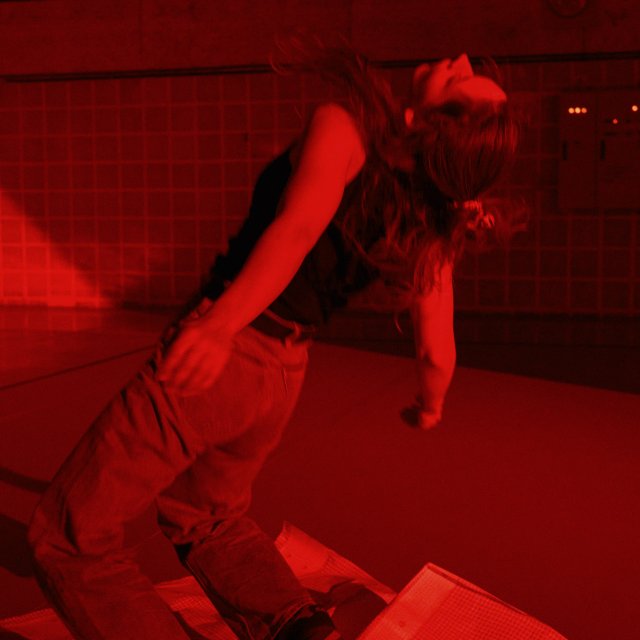 Eine Person bewegt sich auf einer B&uuml;hne unter intensiv rotem Licht. Die Person kniet auf einer gefalteten Gitterplane, der Oberk&ouml;rper ist weit zur&uuml;ckgelehnt. Im Hintergrund sind gekachelte Wandfl&auml;chen und technische Installationen sichtbar.