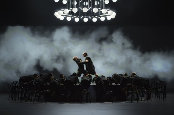 HSB: Nachtträume (c) Sinah Osner A large ensemble gathers around a long table on stage. Above the scene hangs a circular chandelier filled with glowing spherical lights. Two people meet at the center while haze drifts across the background.