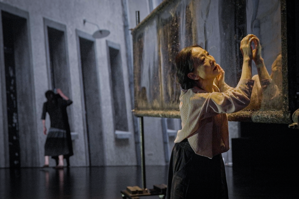 Wen Hui and Eiko Otake in front of a black background. Their bodies intertwined, with concentrated and strained expressions on their faces. The pale light has an oppressive effect