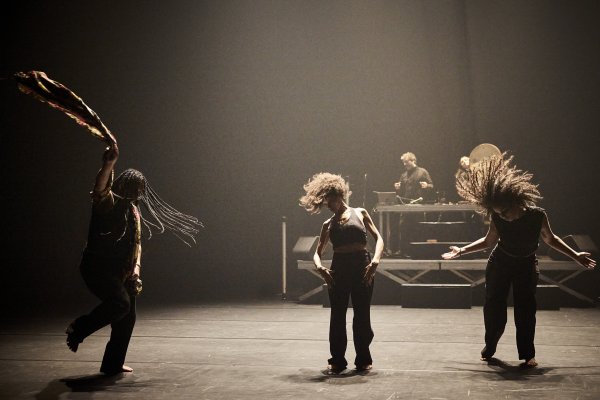 ALI CHAHROUR: WHEN I SAW THE SEA (c) Christophe Raynaud De Lage Three people move across a stage illuminated by warm spotlighting. Dynamic shapes emerge through body positions and hair movement in the foreground. In the background, two additional people work at an elevated sound and music workstation.