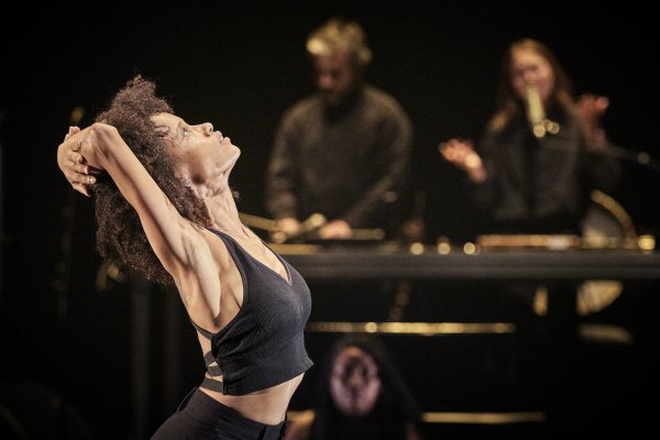 ALI CHAHROUR: WHEN I SAW THE SEA (c) Christophe Raynaud De Lage A performer* stands at the front of a stage with arms folded behind the head and face tilted upward. In the softly blurred background, additional people are positioned at an elevated music and technical workstation. Warm stage lighting contrasts with the dark surroundings.