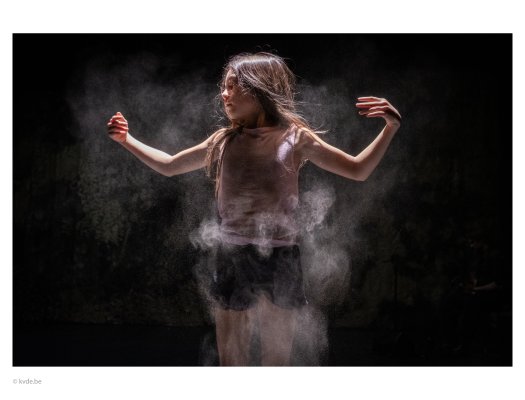 A child stands in a spotlight against a dark background. Fine dust or powder surrounds the body and becomes visible in the light. Raised arms and drifting particles create a striking visual effect.